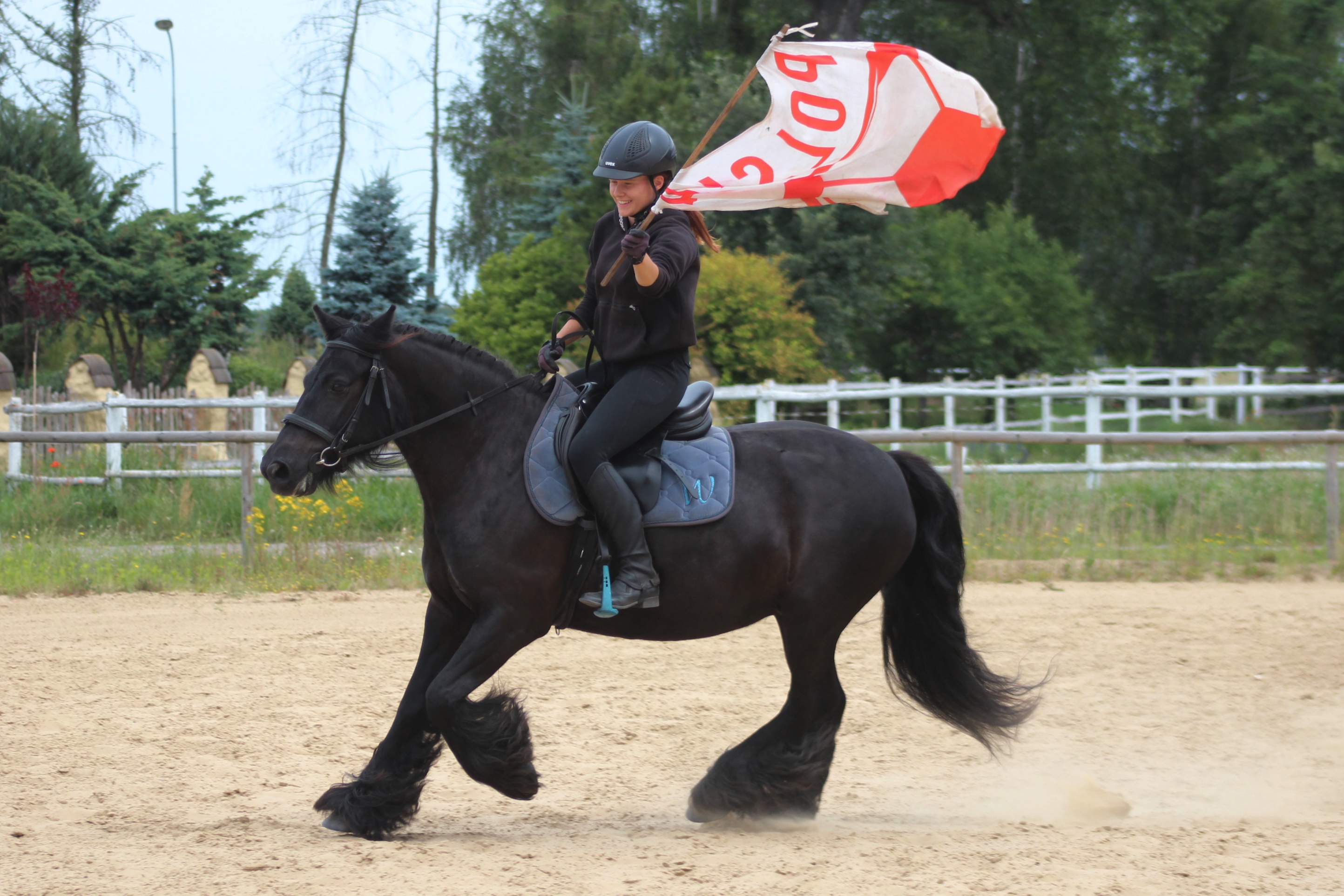 black pony cantering in an arena, the rider carrying a red and white banner